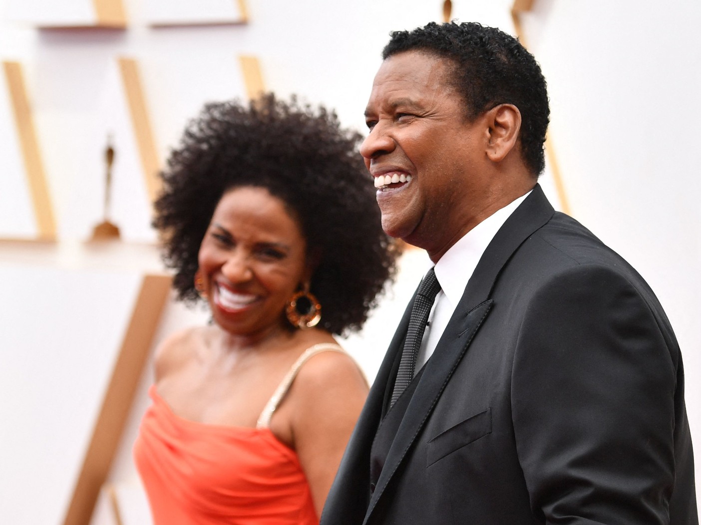 Paulette Washington, in an orange dress, laughs with husband Denzel Washington while walking the red carpet