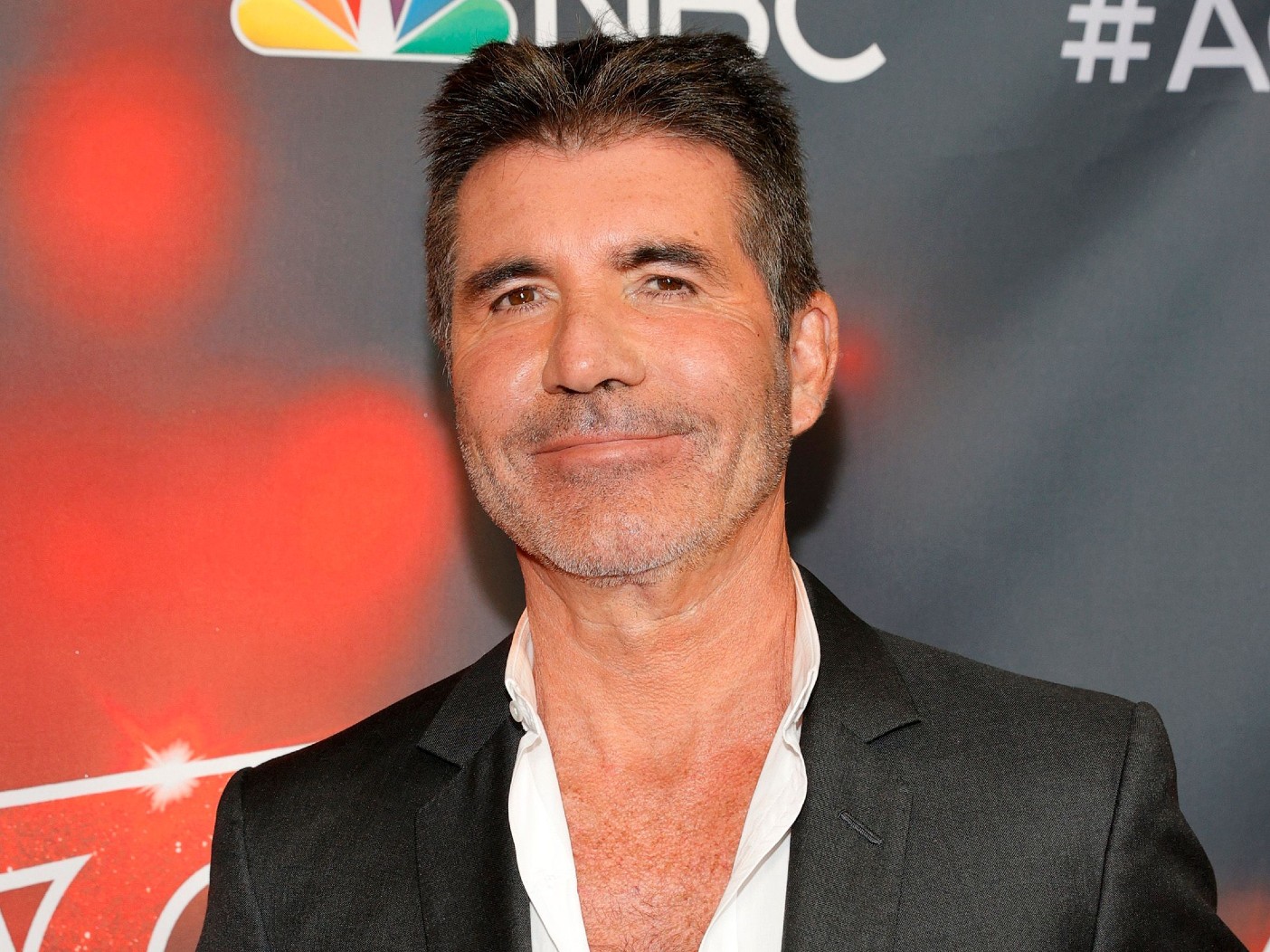 Simon Cowell smiles while wearing a white dress shirt and black blazer
