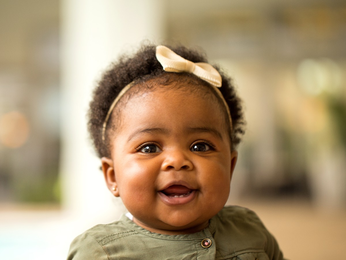 A baby smiles while wearing a forest green outfit and a pale bow headband