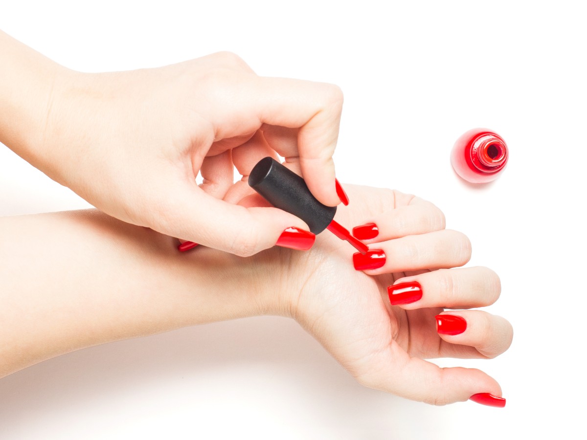 pair of hands in frame, one painting the other's fingernails a bright red color. There is a bottle of red nail polish in frame. All of this is happening on a stark white backdrop