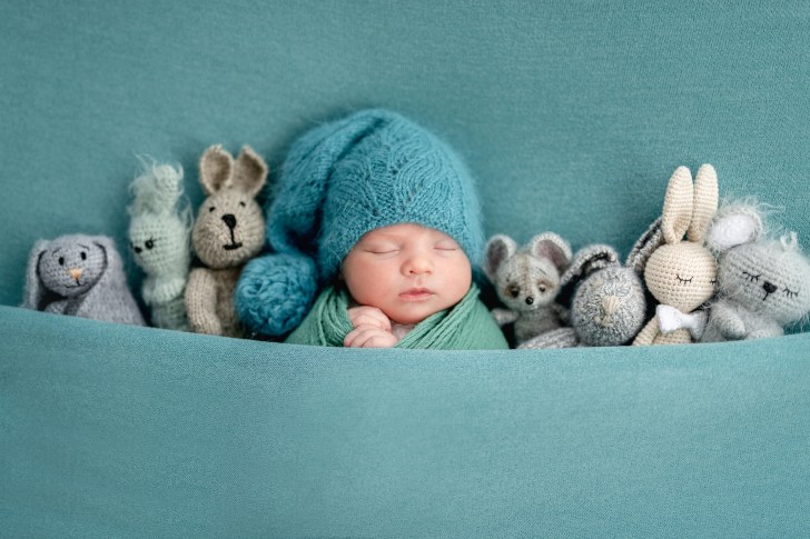 A baby boy sleeps in blue outfit under blue blanket surrounded by tucked-in stuffed animals