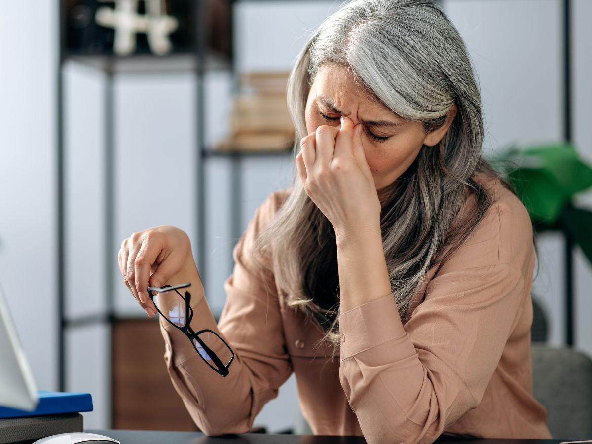 Woman with gray hair sitting at desk pinching brow in stress