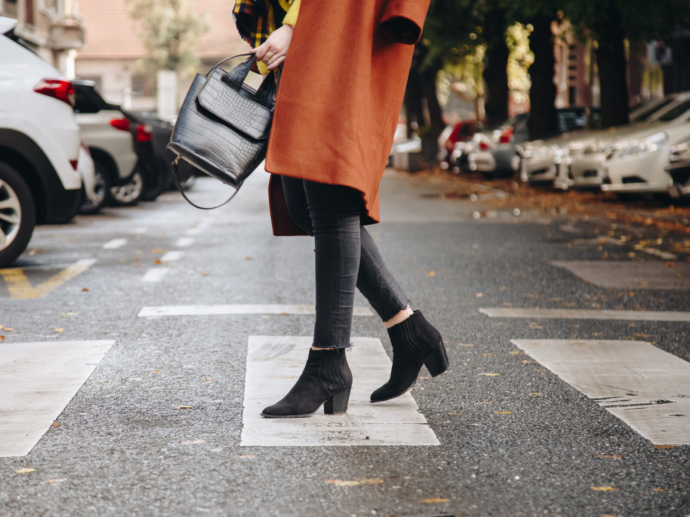 a woman crosses the street wearing black booties and an orange coat