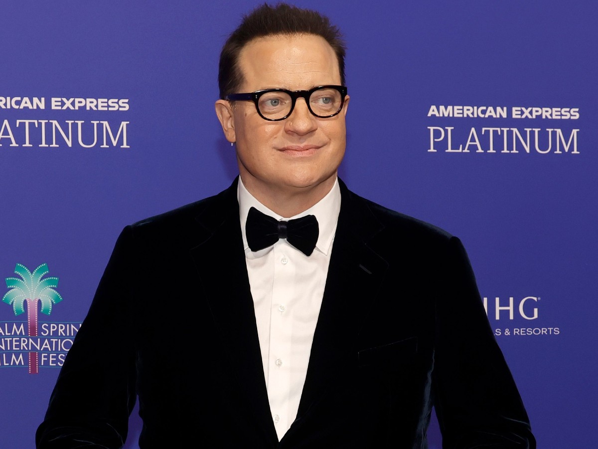 Brendan Fraser smiles in black suit and matching bowtie against blue backdrop