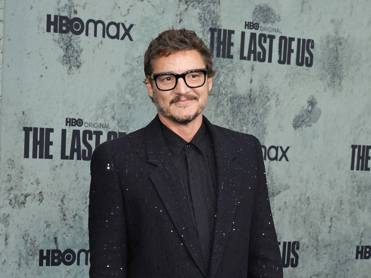 Pedro Pascal smiling in a suave black jacket and suit with black rimmed glasses